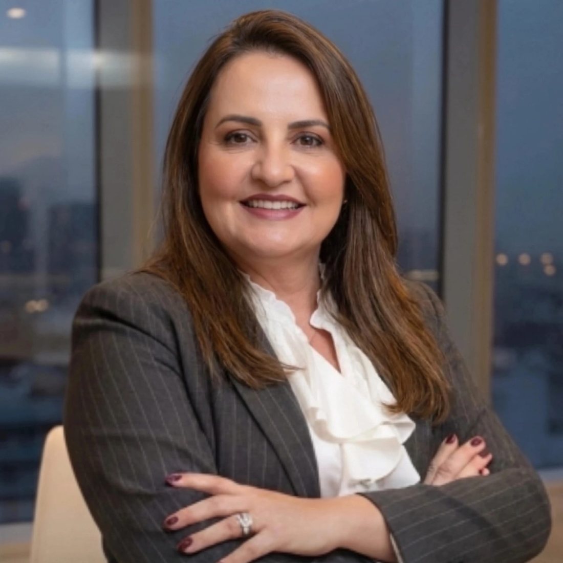 Renata Madrazo de terno risca de giz, braços cruzados, diante de uma janela